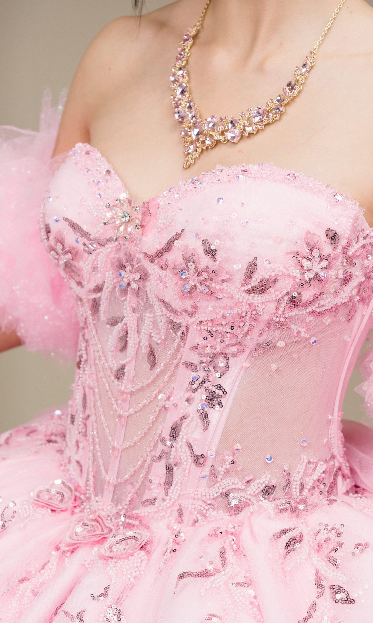 A person wearing the Quinceanera Dress 8380J by Cinderella Couture, a pink strapless gown with beaded embroidery, floral patterns, and a corset bodice, paired with a gold and pink gemstone necklace.